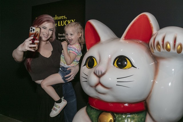 Mother taking a photo with a phone camera of herself and her daughter next to a sculpture of a cat waving